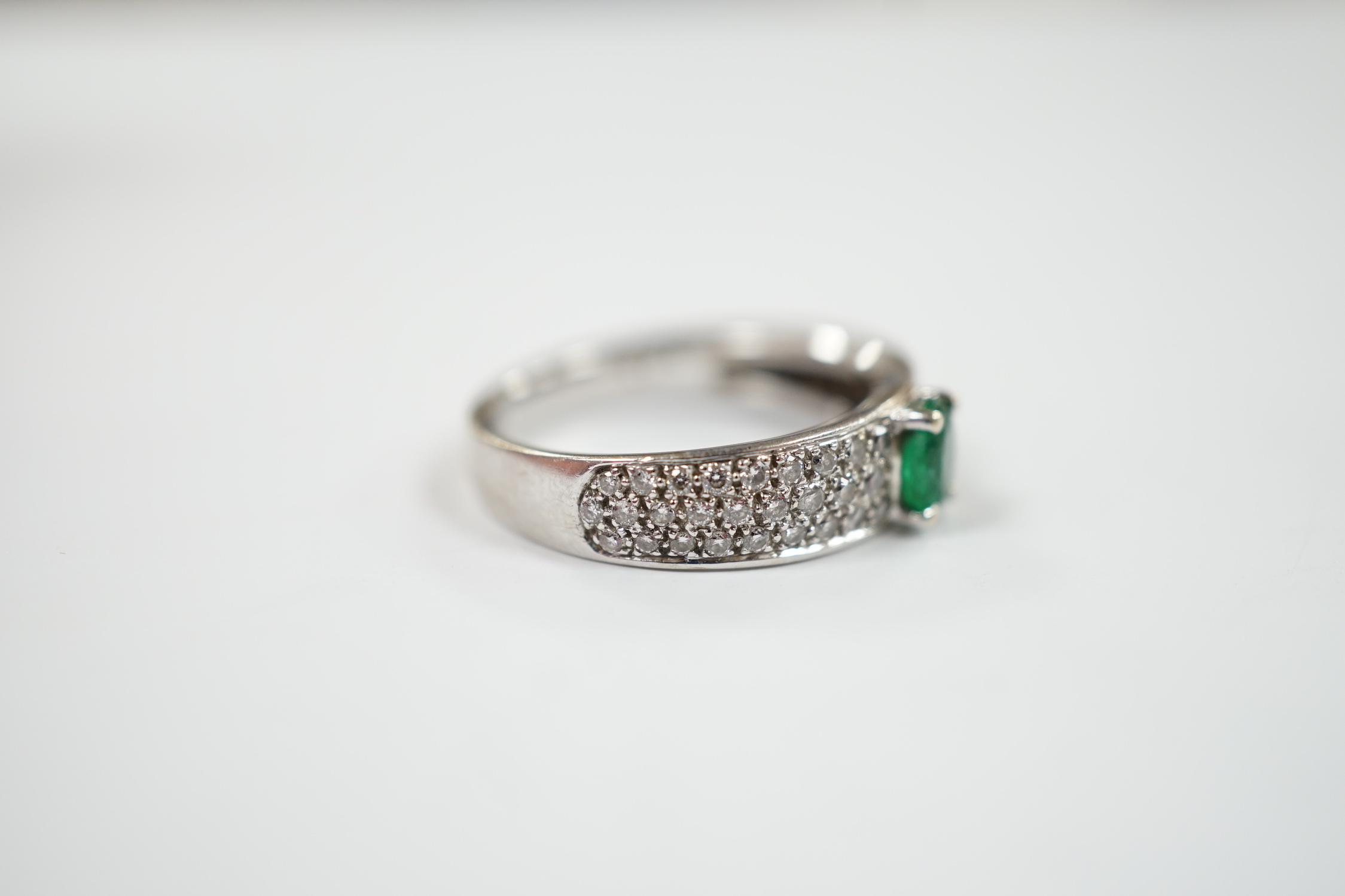 A modern 18ct white gold and single stone oval cut emerald set ring, with pave set diamond shoulders, size L/M, gross weight 4.3 grams.